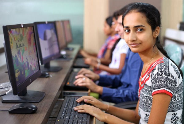 Young woman in a patterned top uses a computer at a desk in a classroom. Others nearby are also focused on monitors. Bright, educational setting.