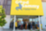People standing outside a store with a yellow "Good Sammy" sign, cutting a ribbon. Clothing racks visible, sunny mood.