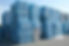 Large stacks of compacted plastic bottles in blue-tinted cubes are piled outdoors on a concrete surface under a clear sky.