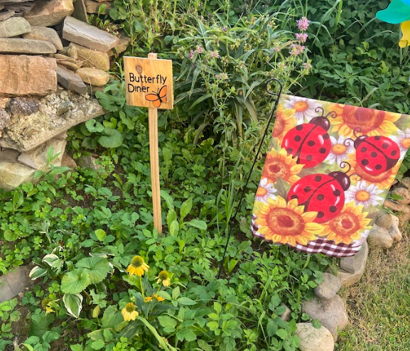 Garden scene with Butterfly Diner sign, ladybug and sunflower flag. Lush green plants, rocks, and colorful flowers in background.