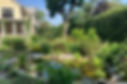 A lush residential garden designed with ecological landscaping principles, featuring native plants, a small pond surrounded by rocks, and a mix of shrubs and perennials near a yellow house. Photo from joegardener®