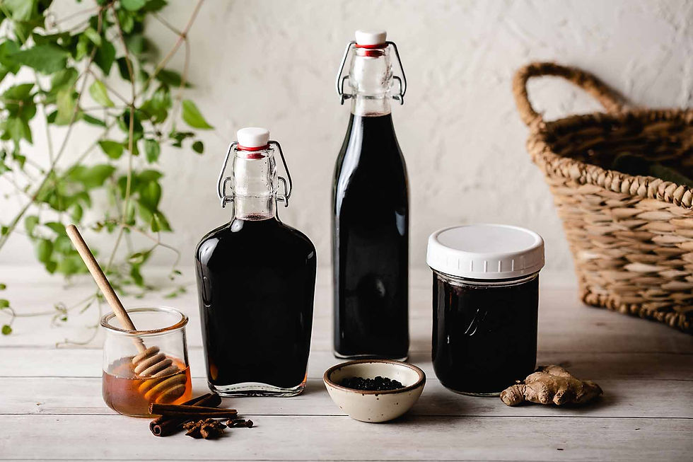 Still-life on a light wooden table featuring two glass flip-top bottles and a screw-top jar filled with deep-purple elderberry elixir, accompanied by a small honey jar with dipper, a bowl of berries, cinnamon sticks, fresh ginger root, a wicker basket, and trailing greenery—showcasing the finished remedies in American Elderberry Alchemy: Cultivating Sambucus canadensis on Your Homestead.