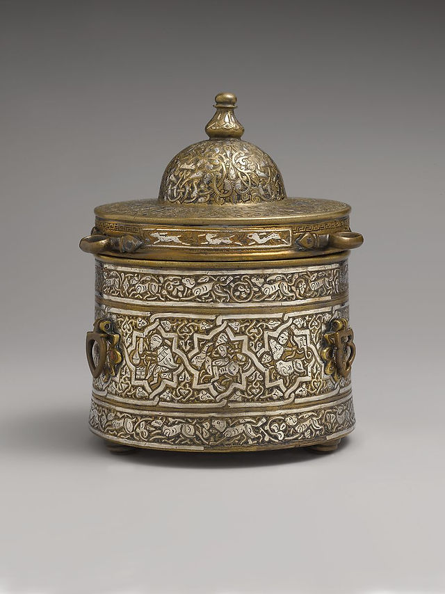Early 13th Century. Iran. Brass; cast, inlaid with silver, copper, and black compound. Inscription in naskhi script: On lid: Continuing glory, increasing prosperity, secure life, and good fortune On top: Glory, prosperity, dominion, dominion .... H. 5 7/8 in. (14.9 cm) H. w/o lid: 3 5/8 in. (9.2 cm) Diam. 4 9/16 in. (11.6 cm). Accession no. 59.69.2a, b.