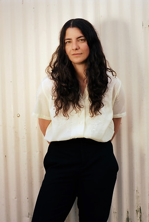 This is a photo of Caitlin Conlen wearing a white shirt and black pants standing against a cream colored background.