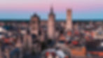 Aerial view of a European city with Gothic architecture, including tall spires and towers at sunset. The sky is pink and blue.