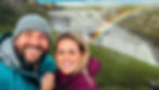 Smiling couple in front of a waterfall with a rainbow. They're outdoors, wearing jackets, with lush green hills in the background.