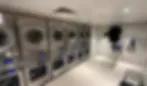 Laundromat with stainless steel washers and dryers, navy uniform hanging, iron on shelf. Brightly lit, tidy, with tiled floor and overhead vents.