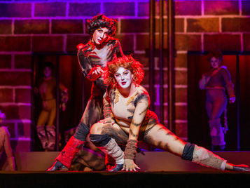Two performers from the musical "Cats" pose together on stage with a red brick background.