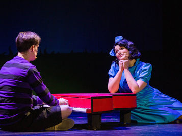 Two actors portraying Lucy and Schroeder in "You're a Good Man Charlie Brown" sitting on stage with a red piano.