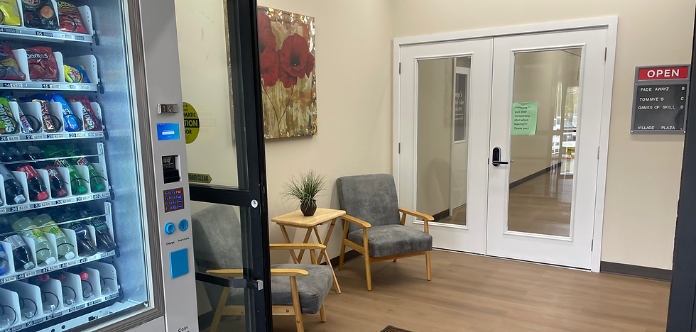 Lobby featuring a vending machine, two gray chairs with a small table in the middle, and a painting above the table of red roses. Lobby has doors that lead to a hallway.