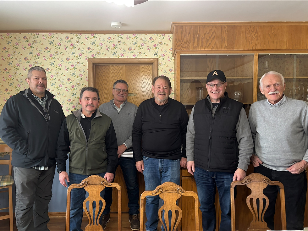 JDI Contracts CEO Roger Hoyum and mining professionals stand smiling in a room with floral wallpaper and wooden cabinets. The mood is cheerful and relaxed.