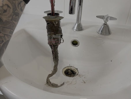 Detailed close-up of a plumber's hands pulling a metal drain insert upward from a white porcelain vanity, revealing a thick, wet buildup of grime and hair clogging the mechanism.