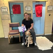 A lady sat with her spaniel between her knees holding a puppy training certificate and rosette