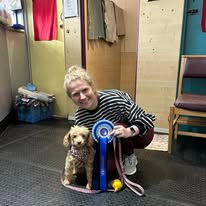 A lady sat next to her poodle dog holding a rosette after passing a training level at Guns and Noses