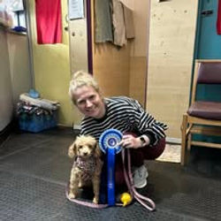 A lady sat next to her poodle dog holding a rosette after passing a training level at Guns and Noses