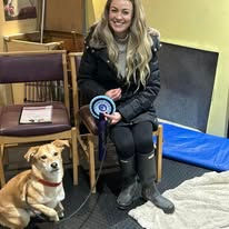 A lady sat next to her cross breed dog holding a rosette after passing a training level at Guns and Noses