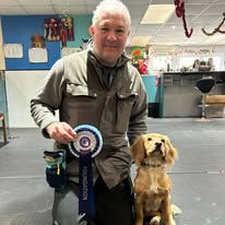 A gentleman sat next to his spaniel dog holding a rosette after passing a training level at Guns and Noses