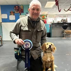 A gentleman sat next to his spaniel dog holding a rosette after passing a training level at Guns and Noses