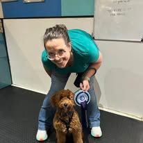 A lady sat next to her cockerpoo dog holding a rosette after passing a training level at Guns and Noses