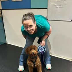 A lady sat next to her cockerpoo dog holding a rosette after passing a training level at Guns and Noses
