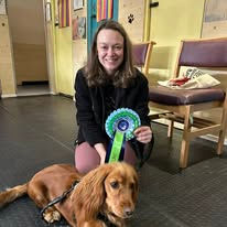 A lady sat next to her spaniel dog holding a rosette after passing a training level at Guns and Noses