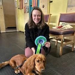 A lady sat next to her spaniel dog holding a rosette after passing a training level at Guns and Noses