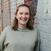 Anna Bennecke lacht in die Kamera. Sie hat helle Haut, blaue Augen und braune, schulterlange Haare, die oben zurückgenommen sind. Das Porträt zeigt sie von Kopf bis etwa zur Hüfte. Sie trägt kleine Creolen, mehrere Ohrstecker, ein kleines Nasenpiercing und eine feine silberne Kette mit rundem Anhänger. Zu ihrem lockeren mintgrünen Sweatshirt hat sie den Kopf leicht geneigt. Sie lehnt an einer grau-blau gestrichenen Backsteinwand mit Graffiti. Links ist unscharf eine Gasse zu sehen.