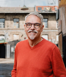 Sven Franke lächelt in die Kamera. Er hat helle Haut, kurze graue Haare, einen grauen Schnurrbart mit hochgezwirbelten Enden, einen kleinen Kinnbart und eine runde schwarze Brille. Das Porträt zeigt ihn von Kopf bis etwa zur Taille, der Oberkörper ist leicht zur Seite gedreht. Er trägt einen orangeroten Pullover mit V-Ausschnitt. Hinter ihm liegt unscharf ein Hinterhof mit einem Altbau und bunt bemalten und beklebten Wänden.