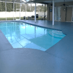 Indoor pool with light blue water, surrounded by a spacious patio. Glass enclosure and trees visible outside. Calm and serene atmosphere.