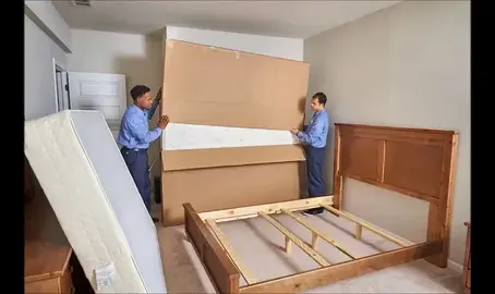 Two movers carrying a large boxed mattress in a bedroom with a wooden bed frame and another mattress leaning against the wall.