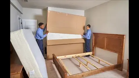 Two movers carrying a large boxed mattress in a bedroom with a wooden bed frame and another mattress leaning against the wall.