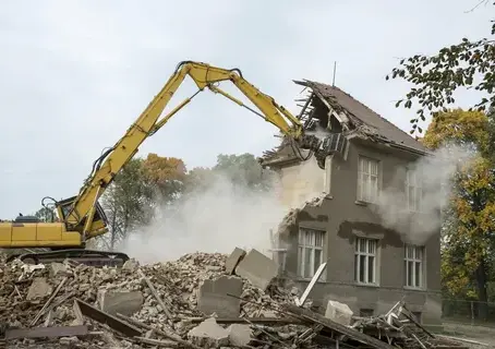 Excavator demolishing an old two-story building with debris around.