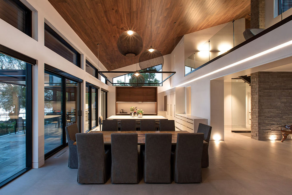 Modern dining room with long wood table, gray chairs, kitchen island, warm wood ceiling, large windows, and stone wall at night. A lighting scene can provide comfortable illumination around the dining area while keeping surrounding spaces softer and drawing attention to architectural details.