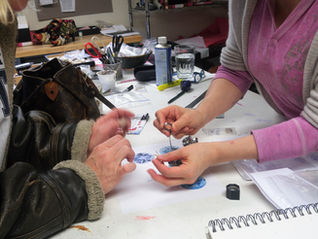 Two people crafting jewelry at a cluttered desk with tools, notes, and a leather bag. One wears a pink sweater, the other a brown jacket.
