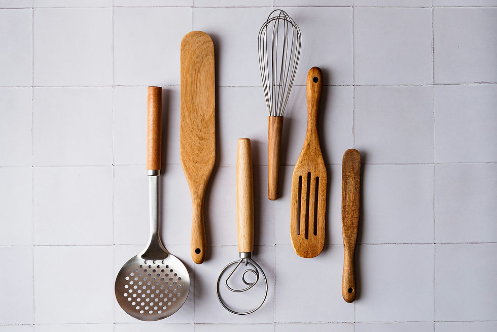 Assorted wooden and metal kitchen utensils laid neatly on white tiled surface, including a whisk, spatula, slotted spoon, and masher.