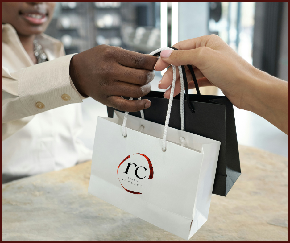 Hands exchange white and black shopping bags. "rc studio jewelry" text is visible. Indoor setting with blurred background.