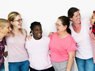 Six women smiling and embracing, wearing pink and casual outfits. Bright, joyful mood against a plain white background.