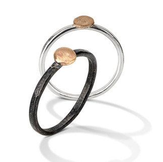 Two rings standing up, one with black finish, the other bright silver; both rings have one gold hammered disk on top.