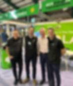 Four men smiling at a solar panel booth with a green "ecoEnergy Improvements" sign. Bright, upbeat atmosphere in an indoor setting.