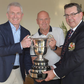 Three men smile while holding a large silver trophy in an indoor setting. One man wears a dark blazer with a crest. Mood is celebratory.