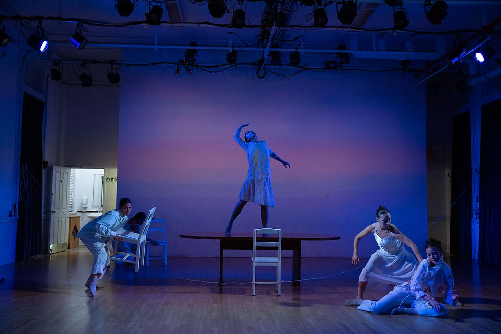 Performers in white costumes dance under blue lighting on a wooden stage. Some stand on tables and chairs, creating dynamic poses.