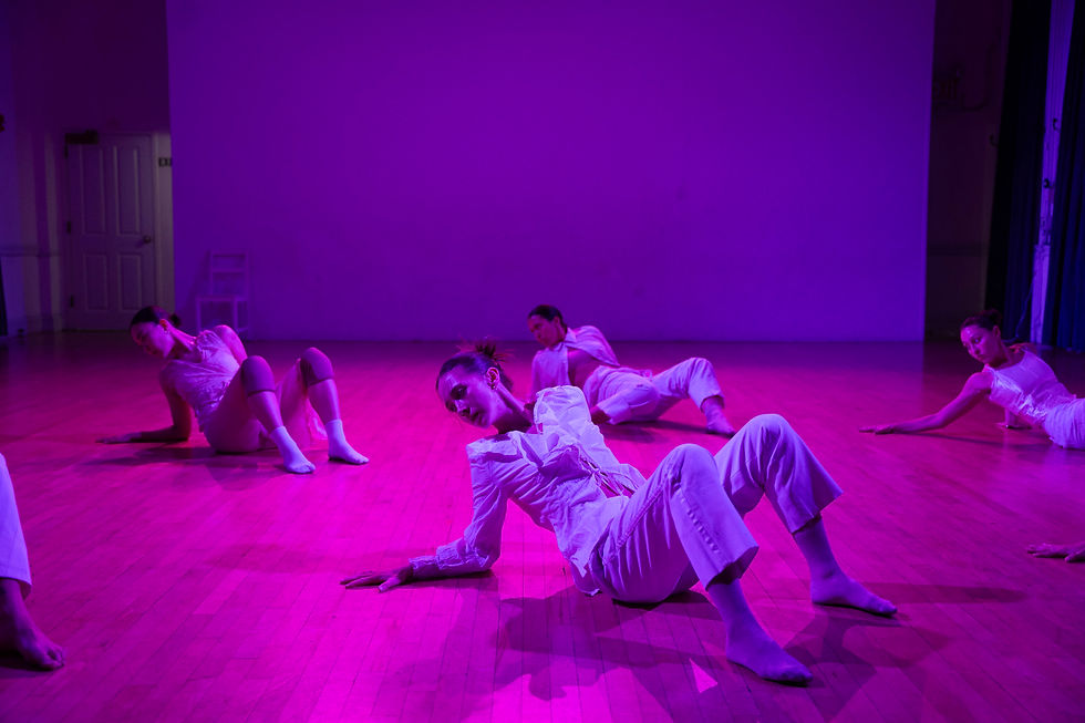 Dancers in white perform on a wooden floor under purple lighting. They are partially reclined, creating a calm, artistic atmosphere.