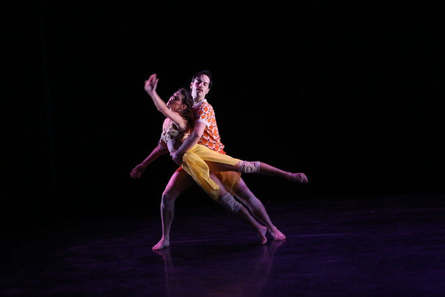 Two dancers in a dramatic lift on a dark stage, lit purple; woman in a yellow skirt leans forward, man in an orange patterned shirt supports her.