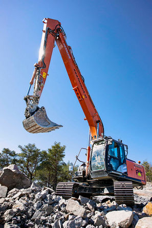 En orange grävmaskin från FMT Mark sträcker sig högt upp mot en klarblå himmel, med skopan öppen och redo att gräva bland sten och rasmassor, vilket visar företagets kapacitet för stora grävarbeten.