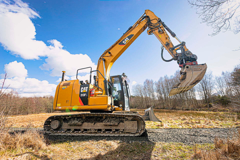 En gul Caterpillar 313F grävmaskin från FMT Mark med skopan lyft i en naturskön miljö med blå himmel och skog i bakgrunden, redo att utföra jordförflyttningsarbete.
