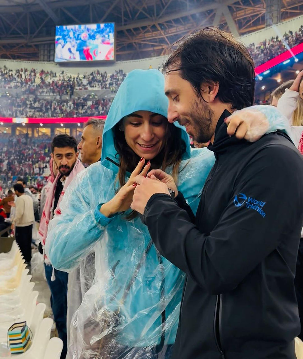 Engagement moment in a stadium, couple in rain ponchos, vibrant crowd in background, portrait, focus on hands and ring