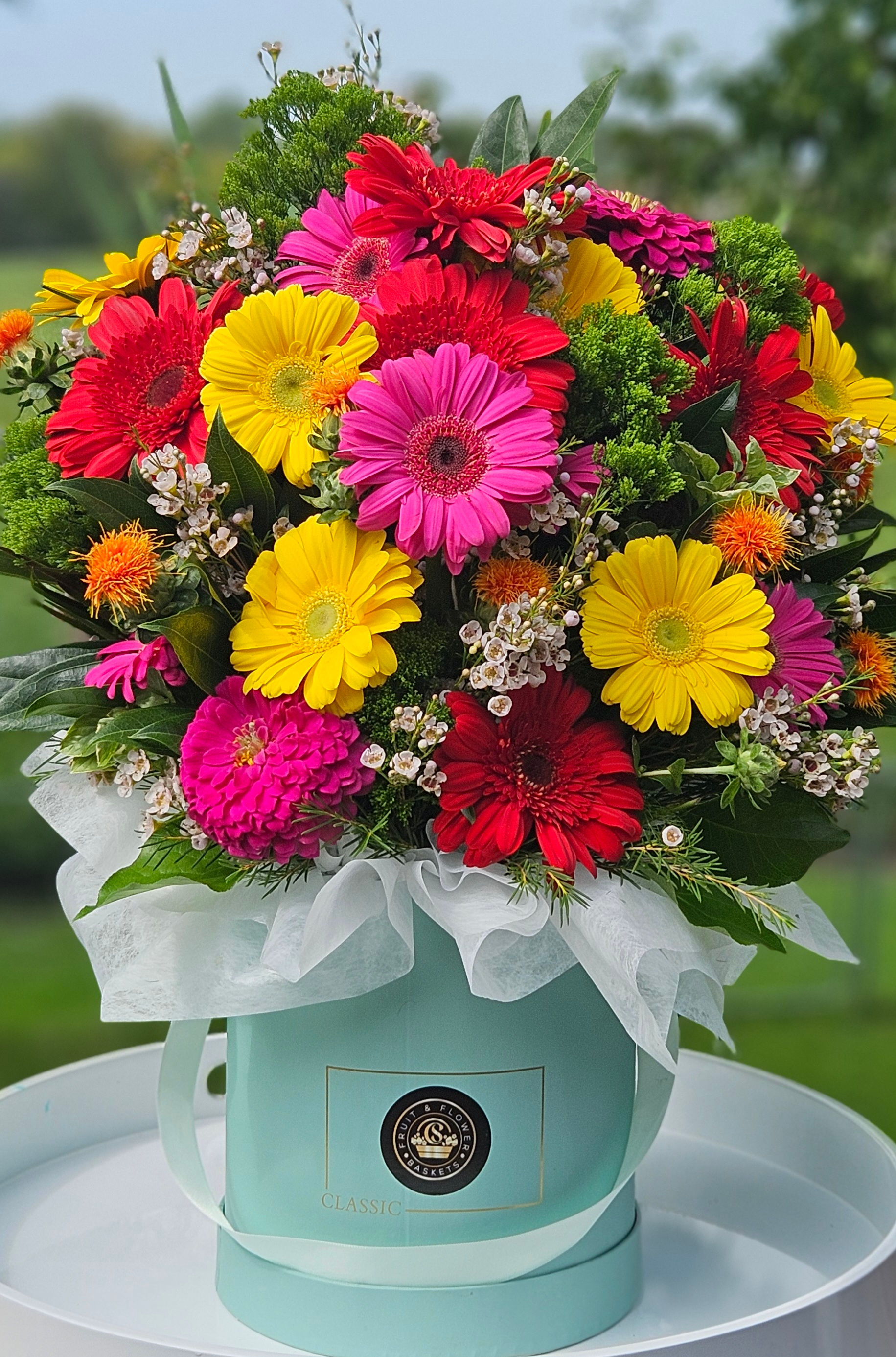 A bouquet of colorful flowers including red, yellow, and pink gerbera daisies arranged in a light blue hat box