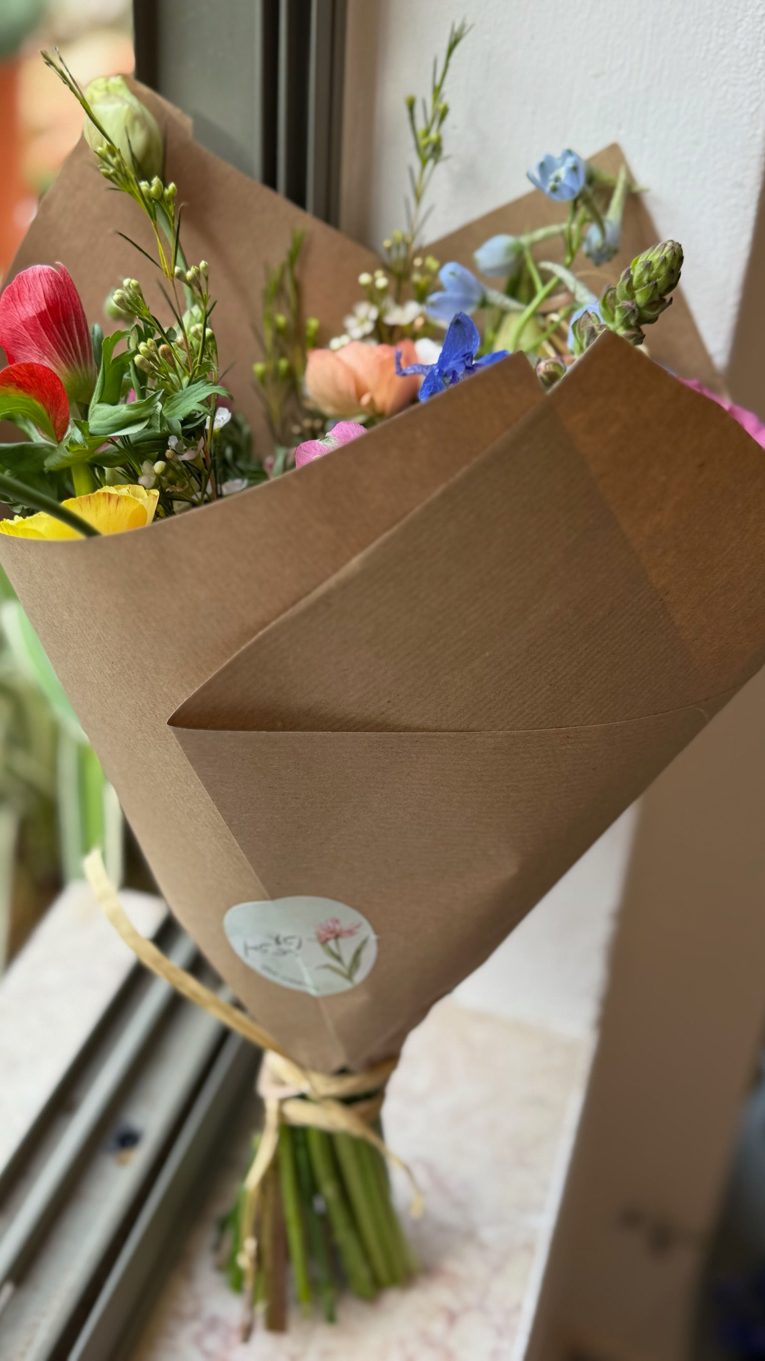 זר פרחים צבעוני, עטוף בנייר חום טבעי וקשור בחוט קש - רשת לול, מונח על אדן חלון