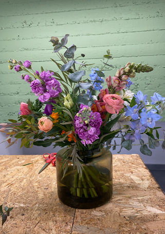 זר פרחי אביב גדול וגבוה בתוך כלי זכוכית כהה ירוק, המשלב גווני סגול, ורוד ותכלת עם פרחי מתיולה (סטוק) ודלפיניום.