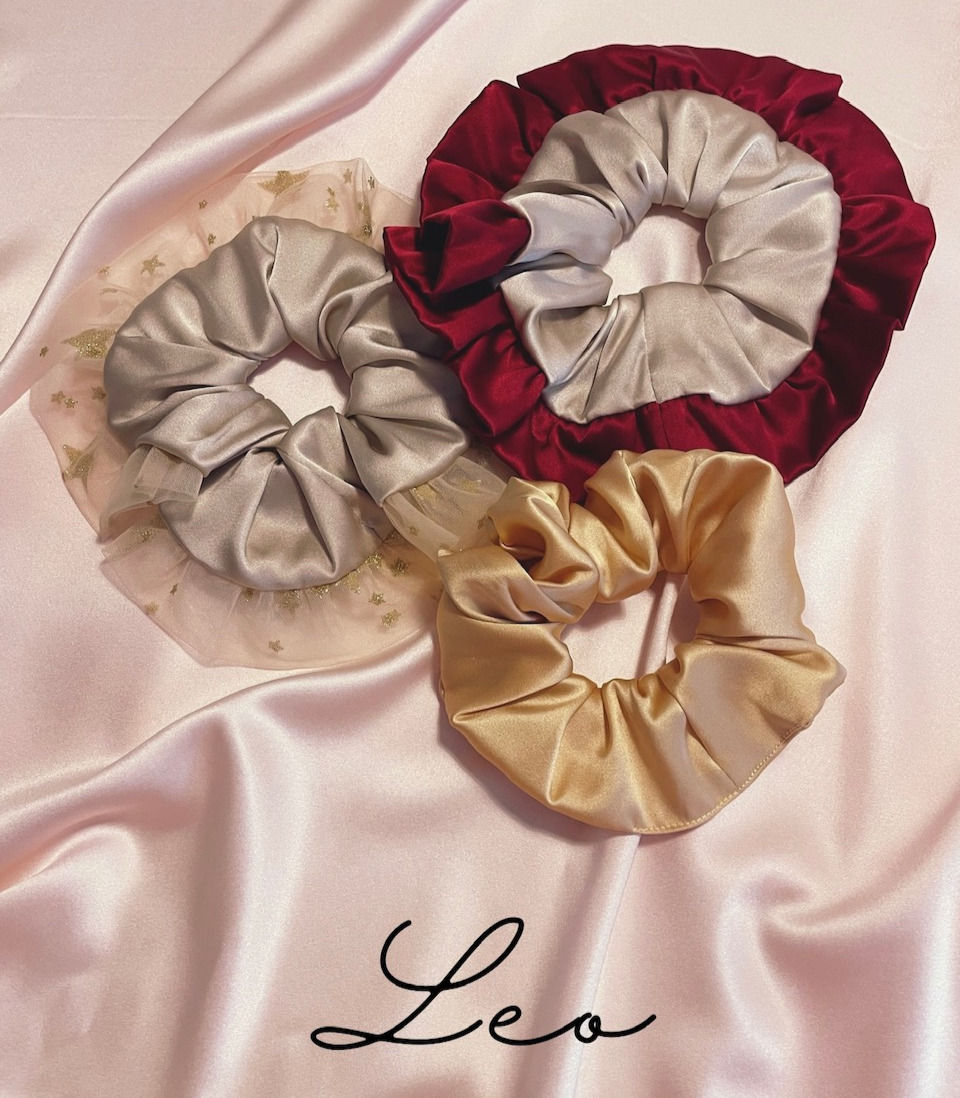 a large double ruffle silk scrunchie in golden beige with deep red outer ruffle, a second double ruffle scrunchie in golden beige with a glitter star tulle outer ruffle and a gold silk scrunchie against a pale pink silk background with Leo text